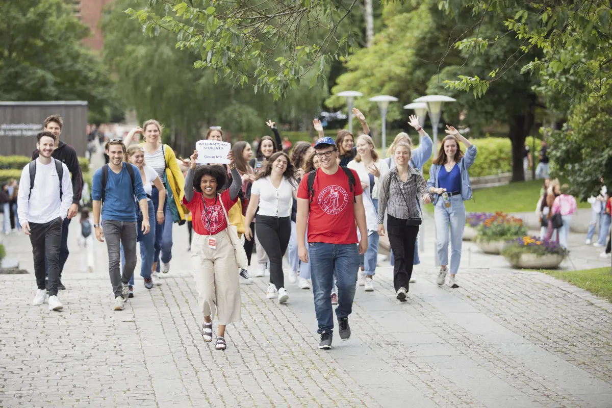 UiO studenter kommer g?ende side og side opp trappen mot fontenen p? Blindern. To faddere g?r fremst i r?de t-skjorter med UiO-logo p?. Den ne fadderen holder opp et skilt med med p?skriften: Internasjonale studenter. 